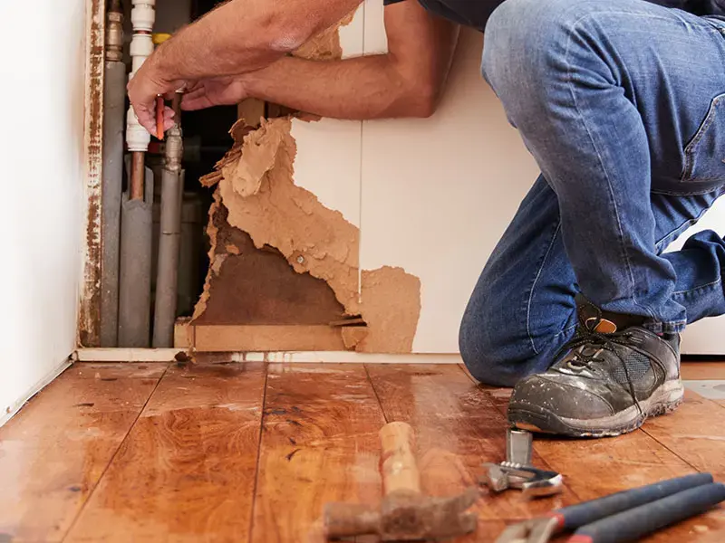 Drywall opened at the base of a wall to reach the supply pipes, with water on the wood floor